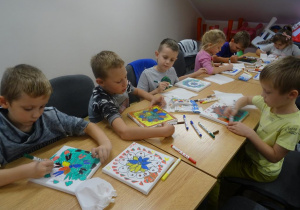 Grupa dzieci siedzi przy stole, trzymają w ręku mazaki i dokańczają malowanie na płótnie.
